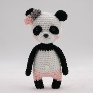 Könnte beinhalten: Ein weißer und schwarzer gehäkelter Pandabär mit rosa Akzenten. Der Panda hat eine rosa Blume auf dem Kopf und schwarze Augen.