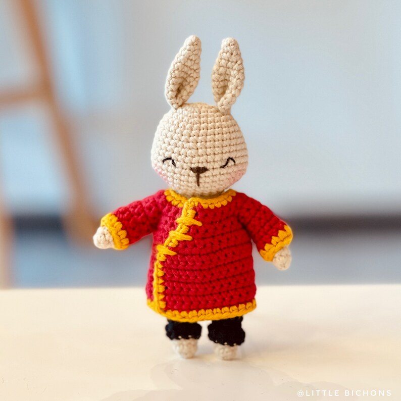 May include: A crocheted bunny wearing a red and yellow Chinese-style jacket. The bunny has a white face with black eyes and a black nose.