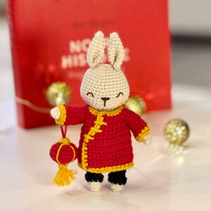 May include: A crocheted white rabbit wearing a red and yellow Chinese-style outfit and holding a red and yellow lantern.