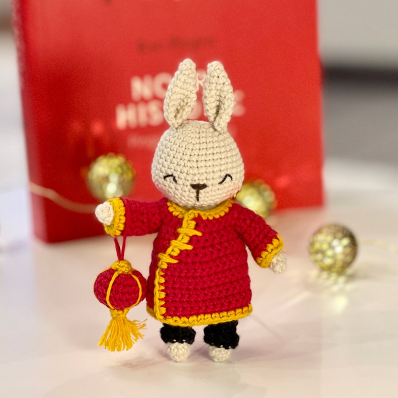 May include: A crocheted white rabbit wearing a red and yellow Chinese-style outfit and holding a red and yellow lantern.
