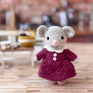 Pode incluir: Uma boneca de rato em crochê com fio cinza claro, olhos de contas pretas e bochechas rosadas. O rato veste um vestido magenta escuro com uma gola branca recortada e dois pequenos botões brancos na frente, além de orelhas arredondadas.