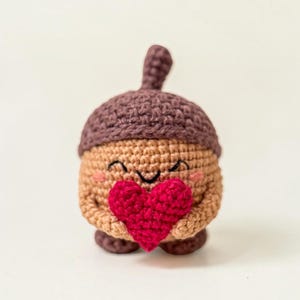 Könnte beinhalten: Eine kleine gehäkelte Eichelfigur mit einer dunkelbraunen Kappe und einem hellbraunen Körper. Die Eichel hat aufgestickte schwarze Augen und ein Lächeln, rosa Wangen und hält ein leuchtend rotes, strukturiertes, gehäkeltes Herz in den Armen.