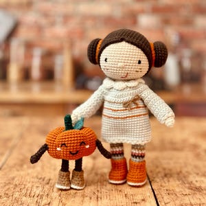 May include: A crocheted doll with brown hair and a blue and orange striped dress holds a crocheted pumpkin with a smiling face.