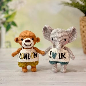 Puede incluir: Dos animales de peluche tejidos a crochet, un oso marrón y un elefante gris, que llevan camisas blancas con texto bordado. La camisa del oso dice "LONDON" y la camisa del elefante dice "I ♥ UK".
