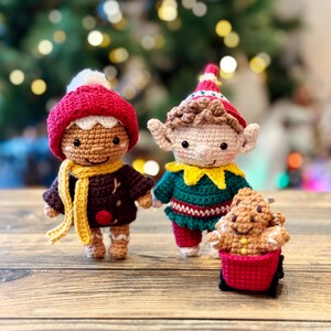 Peut inclure: Deux décorations d'elfes de Noël en crochet. Un elfe porte un chapeau rouge et blanc et une écharpe brune. L'autre elfe porte un chapeau vert et rouge et une écharpe jaune. L'elfe de droite tient une décoration en pain d'épices.