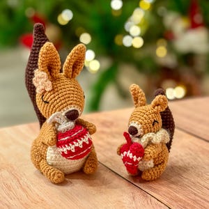 Peut inclure: Deux figurines de crochet en forme d'écureuil. L'un tient une boule de Noël rouge et blanche, l'autre un gland rouge. Les écureuils ont une fourrure brune, un ventre blanc et une queue marron foncé. L'arrière-plan est flou avec des lumières de Noël.