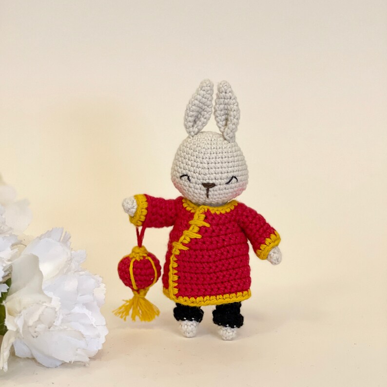 May include: A white crocheted bunny wearing a red and yellow Chinese-style robe and holding a red and yellow lantern.