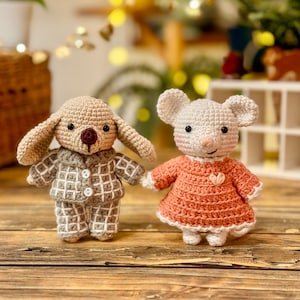 May include: Two crocheted stuffed animals, a dog and a mouse, wearing pajamas. The dog is wearing a gray and white plaid pajama set and the mouse is wearing a pink dress with a white heart on the front.