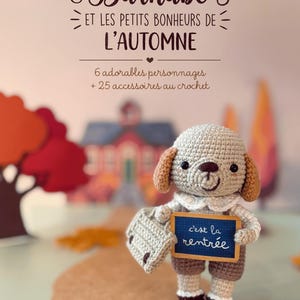 May include: A crochet dog figurine holding a small briefcase and a sign that reads "c'est la rentrée". The book cover features the title "Barnabé et les petits bonheurs de l'automne" with the subtitle "6 adorables personnages + 25 accessoires au crochet".