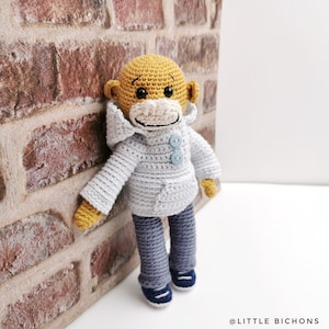 Puede incluir: Un mono de crochet que lleva una chaqueta con capucha azul claro y pantalones vaqueros azules. El mono tiene la cara amarilla y los ojos marrones.