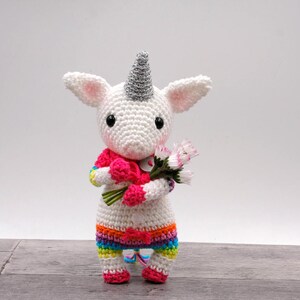 Peut inclure: Un jouet en crochet en forme de licorne blanche tenant un bouquet de fleurs roses. La licorne porte une jupe rayée arc-en-ciel et une écharpe rose.