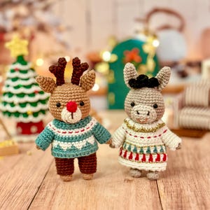 以下が含まれることがあります： 2つの編みぐるみのクリスマスオーナメント：茶色の角と赤い鼻を持つトナカイとロバ。トナカイは白い模様の入ったティール色のセーターを着ており、ロバは赤と金のアクセントの白いドレスを着ています。背景には編みぐるみのクリスマスツリーがあります。