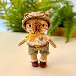 Op de afbeelding: Handgemaakte gehaakte wombatpop met een kaki safarihoed, korte broek en een crèmekleurig shirt met een gele sjaal. De pop heeft een bruine neus, roze wangen en bruine laarzen. De pop is ongeveer 15 cm hoog.