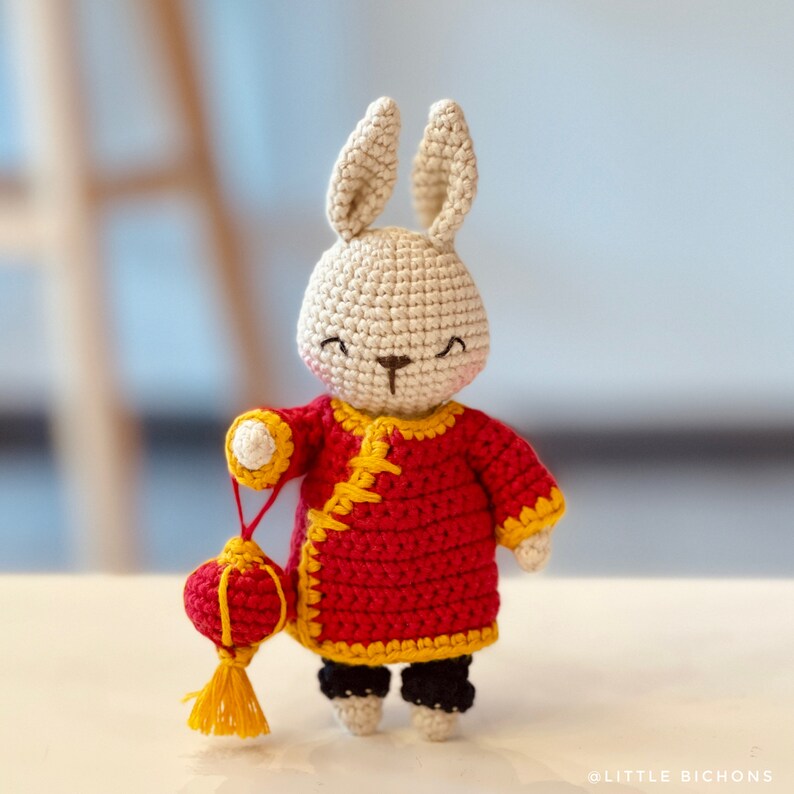 May include: A crocheted rabbit wearing a red and yellow Chinese-style outfit and holding a red and yellow lantern.