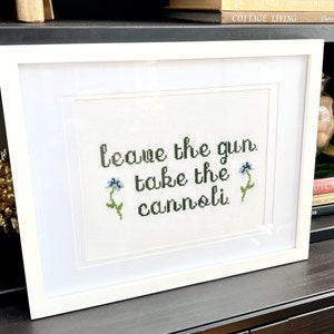 May include: White framed cross-stitch embroidery with the text "Leave the gun, take the cannoli" and two blue flowers.