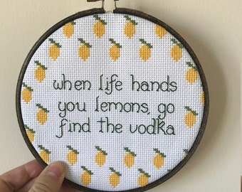 May include: A white embroidery hoop with a brown wooden frame. The embroidery features a white background with yellow lemons and green leaves. The text "when life hands you lemons, go find the vodka" is embroidered in green thread.