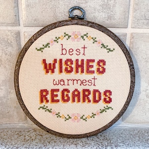 Może przedstawiać: Biały tamborek do haftu z brązową drewnianą ramą. Haft przedstawia wzór kwiatowy z tekstem "best wishes warmest regards" w kolorze czerwonym i żółtym.