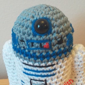 Könnte beinhalten: Ein gehäkeltes R2-D2 Spielzeug mit blauen und weißen Streifen, einem schwarzen Auge und einem roten Mund. Das Spielzeug steht auf einer Holzoberfläche und hat den Text "Handmade with Love... Shoppygifts" auf der Unterseite.
