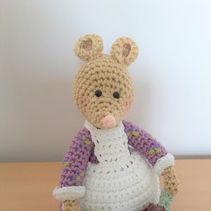 May include: A handmade crochet mouse doll with a tan body, pink nose, and black eyes. It wears a white apron over a purple and green floral patterned dress. The mouse holds a brown pot and a green and tan brush.