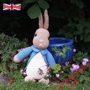Może przedstawiać: Pluszowa zabawka Peter Rabbit wykonana na szydełku, ubrana w niebieską kurtkę. Zabawka siedzi w ogrodzie z zielonymi roślinami i różowymi kwiatami. Tekst "Handmade with Love..." jest widoczny w prawym dolnym rogu obrazu.