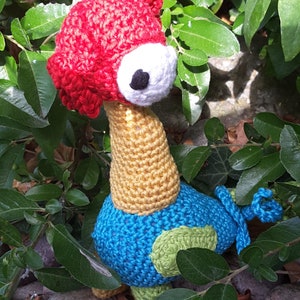 May include: A crocheted toy bird with a red head, yellow neck, blue body, and green accents. The bird has one large white eye with a black pupil.