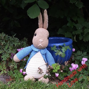 May include: A crocheted stuffed bunny rabbit in a blue jacket, sitting in a garden with green plants and pink flowers. The bunny has brown eyes and a white belly. The text "Handmade with Love..." is visible in the lower right corner of the image.