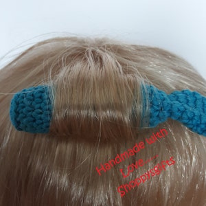 May include: A teal blue crocheted hair tie with a bow on the end. The hair tie is on a person's head and the text "Handmade with Love... Shoppygifts" is written in red below the hair tie.