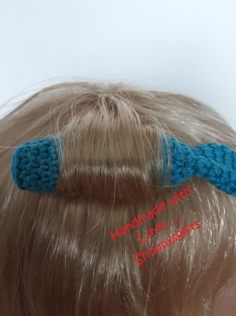 May include: A teal blue crocheted hair tie with a bow on the end. The hair tie is on a person's head and the text "Handmade with Love... Shoppygifts" is written in red below the hair tie.