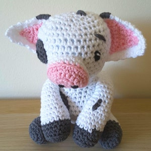 May include: A crocheted white cow plush toy with pink ears, a pink nose, and black eyes. The cow has gray hooves and black spots.
