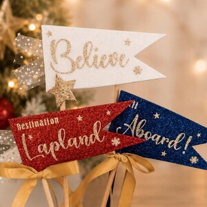 May include: Three glittery pennant flags on wooden sticks. The white flag reads Believe, the red flag reads Destination Lapland, and the blue flag reads All Aboard. Each flag is decorated with gold ribbon bows and sheer star-patterned tulle accents.