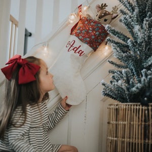 May include: A white Christmas stocking with the name "Ada" in red script, patterned cuff, and a deer toy. The stocking hangs on a white staircase, next to a small Christmas tree. A young girl looks at the stocking.
