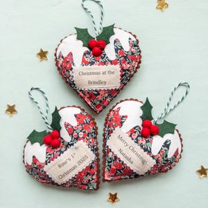Puede incluir: Tres adornos de tela en forma de coraz&oacute;n con estampado floral, partes superiores de tela blanca que simulan glaseado, hojas de acebo de fieltro verde y bayas de pomp&oacute;n rojo. Cada adorno tiene una etiqueta de tela personalizada con el texto Christmas at the Brindley, Rose's 1st Christmas 2025 o Merry Christmas Natasha.