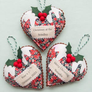Puede incluir: Tres adornos de tela en forma de coraz&oacute;n con estampado floral, glaseado de fieltro blanco, hojas de acebo verdes y bayas rojas. Cada adorno tiene un cordel para colgar y una etiqueta de tela con el texto: Christmas at the Brindley, Rose's 1st Christmas 2025 y Merry Christmas Natasha.