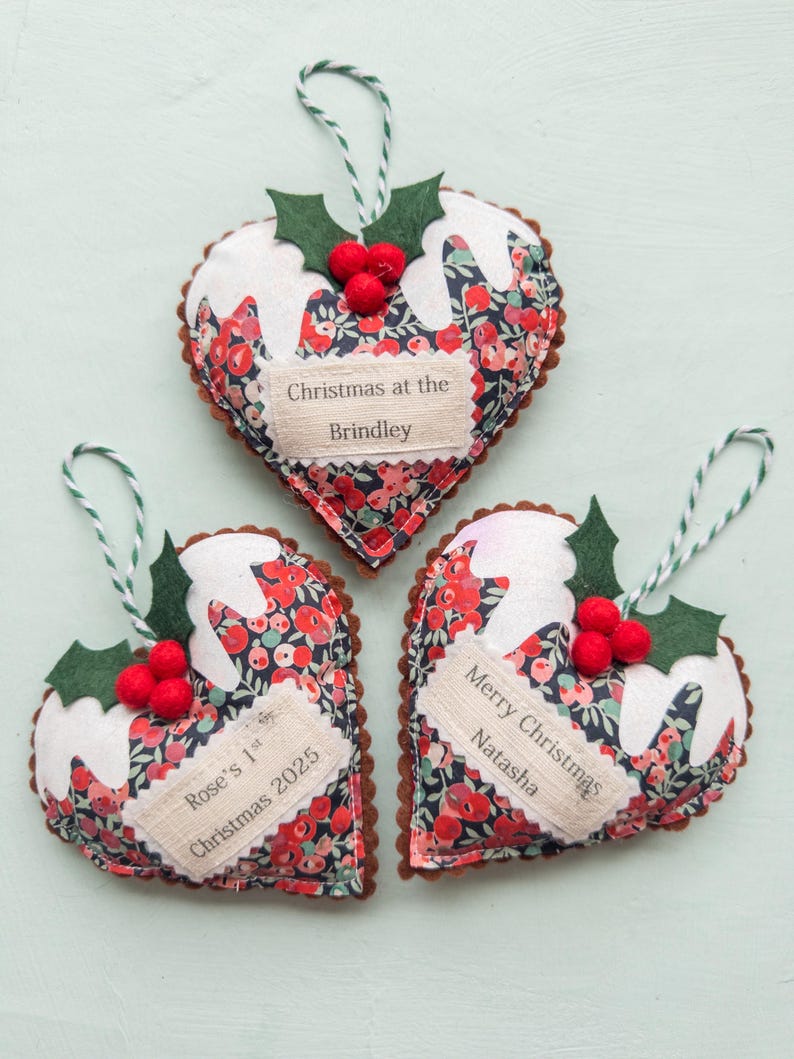 Puede incluir: Tres adornos de tela en forma de coraz&oacute;n con estampado floral, glaseado de fieltro blanco, hojas de acebo verdes y bayas rojas. Cada adorno tiene un cordel para colgar y una etiqueta de tela con el texto: Christmas at the Brindley, Rose's 1st Christmas 2025 y Merry Christmas Natasha.