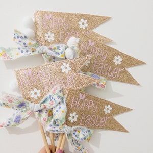 May include: Gold glitter Easter pennants with the text "Hoppy Easter" in pink, decorated with white daisy accents. Each pennant is attached to a wooden stick and decorated with a floral bow and a white bunny tail.