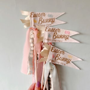 May include: Three pastel pink and white Easter Bunny pennant flags with gold glitter text and decorative ribbons. Each flag reads "Easter Bunny Please Stop Here" with bunny and egg illustrations.
