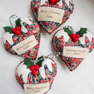 Puede incluir: Cuatro adornos de tela en forma de coraz&oacute;n con un patr&oacute;n floral oscuro, bayas rojas y hojas verdes. Cada adorno tiene glaseado de fieltro blanco, bayas de fieltro rojo y una hoja verde. Las etiquetas dicen Merry Christmas Inma, Sarah, Stefi y Christmas at the Lines.