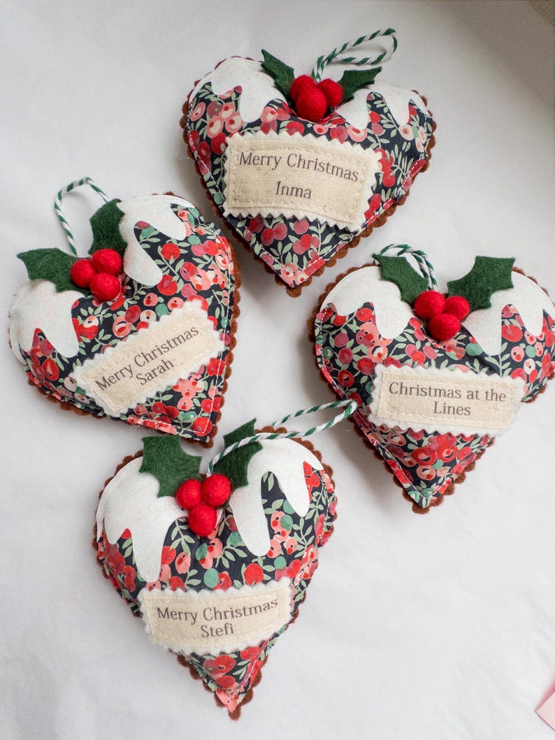Puede incluir: Cuatro adornos de tela en forma de coraz&oacute;n con un patr&oacute;n floral oscuro, bayas rojas y hojas verdes. Cada adorno tiene glaseado de fieltro blanco, bayas de fieltro rojo y una hoja verde. Las etiquetas dicen Merry Christmas Inma, Sarah, Stefi y Christmas at the Lines.