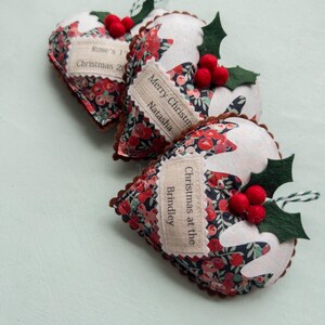 Puede incluir: Tres adornos de tela en forma de coraz&oacute;n con un estampado floral en rojo, azul y verde. Cada adorno tiene una parte superior de fieltro blanco, bayas de fieltro rojo y hojas de acebo de fieltro verde. Una etiqueta de tela crema tiene texto.