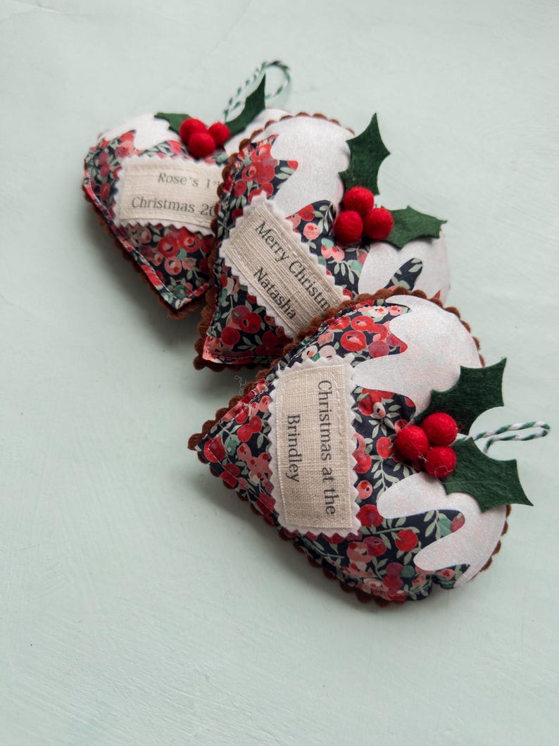 Puede incluir: Tres adornos de tela en forma de coraz&oacute;n con un estampado floral en rojo, azul y verde. Cada adorno tiene una parte superior de fieltro blanco, bayas de fieltro rojo y hojas de acebo de fieltro verde. Una etiqueta de tela crema tiene texto.