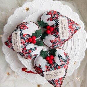 Puede incluir: Tres adornos navide&ntilde;os de tela con forma de pudin, estampado floral oscuro, glaseado de fieltro blanco, hojas de acebo verdes y bayas rojas. Cada pieza tiene una etiqueta de tela beige con el texto: Merry Christmas Sarah, Christmas at the Lines y Merry Christmas Stefi.
