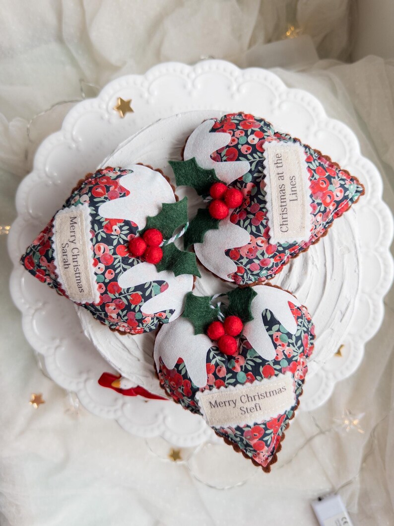 Puede incluir: Tres adornos navide&ntilde;os de tela con forma de pudin, estampado floral oscuro, glaseado de fieltro blanco, hojas de acebo verdes y bayas rojas. Cada pieza tiene una etiqueta de tela beige con el texto: Merry Christmas Sarah, Christmas at the Lines y Merry Christmas Stefi.