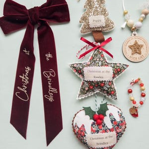 Puede incluir: Una colecci&oacute;n de adornos navide&ntilde;os que incluye un lazo de terciopelo rojo oscuro con texto dorado que dice Christmas at the Brindleys, un &aacute;rbol de purpurina dorada, una estrella de tela floral con texto, un adorno de pudin y una etiqueta de madera.
