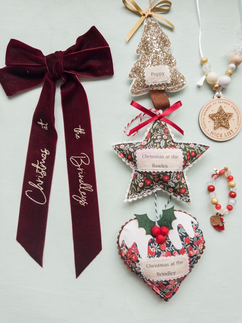 Puede incluir: Una colecci&oacute;n de adornos navide&ntilde;os que incluye un lazo de terciopelo rojo oscuro con texto dorado que dice Christmas at the Brindleys, un &aacute;rbol de purpurina dorada, una estrella de tela floral con texto, un adorno de pudin y una etiqueta de madera.