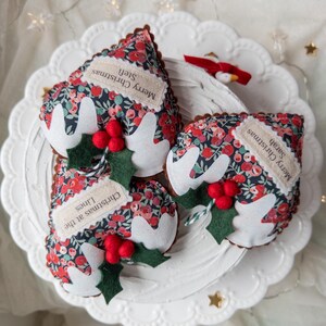 Puede incluir: Tres adornos navide&ntilde;os de tela con forma de pudin, con un estampado floral oscuro de flores rojas y azules. Cada pieza tiene glaseado de fieltro blanco, hojas de acebo de fieltro verde y bayas rojas. Las etiquetas dicen Merry Christmas Stefi, Sarah y Christmas at the Lines.