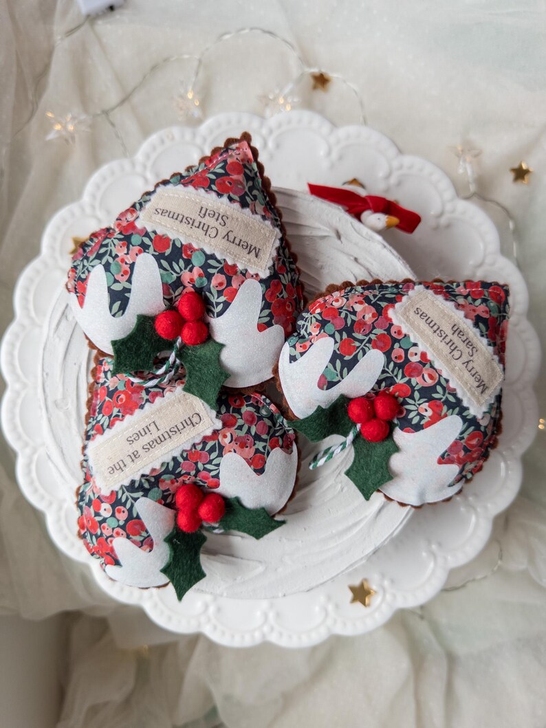 Puede incluir: Tres adornos navide&ntilde;os de tela con forma de pudin, con un estampado floral oscuro de flores rojas y azules. Cada pieza tiene glaseado de fieltro blanco, hojas de acebo de fieltro verde y bayas rojas. Las etiquetas dicen Merry Christmas Stefi, Sarah y Christmas at the Lines.