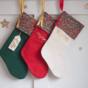 May include: Three Christmas stockings in dark green, red, and off-white fabric. Each features a floral cuff with red and green patterns, gold ribbon hangers, and gold glitter lettering. The green stocking has a tag labelled Ella, while others show names.