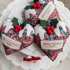 Puede incluir: Tres adornos de tela en forma de coraz&oacute;n con un patr&oacute;n floral oscuro en tonos rojos y azules. Cada adorno est&aacute; rematado con glaseado de fieltro blanco, hojas de acebo de fieltro verde y bayas de fieltro rojas. Etiquetas Merry Christmas Sarah, Stefi y Lines.