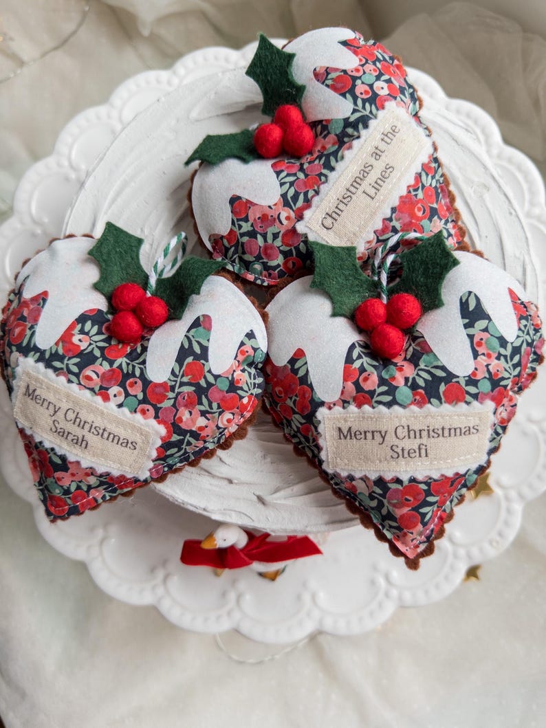 Puede incluir: Tres adornos de tela en forma de coraz&oacute;n con un patr&oacute;n floral oscuro en tonos rojos y azules. Cada adorno est&aacute; rematado con glaseado de fieltro blanco, hojas de acebo de fieltro verde y bayas de fieltro rojas. Etiquetas Merry Christmas Sarah, Stefi y Lines.
