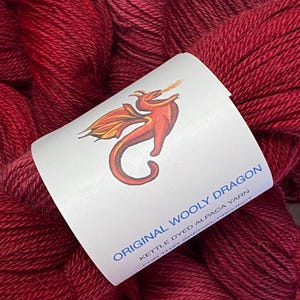 May include: Burgundy-colored skeins of Original Wooly Dragon kettle-dyed alpaca yarn. A white label features a red dragon illustration and text that reads "Original Wooly Dragon" and "For That Special Project."